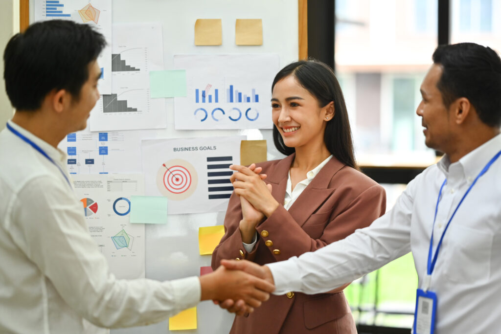 Grateful businesswoman clapping hands while businesspeople shaking hands after successful negotiations at meeting.