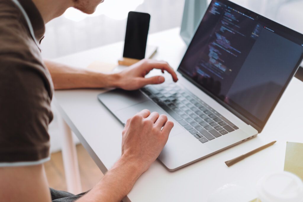 Young man mobile developer, programmer writes program code on a laptop computer in home office.